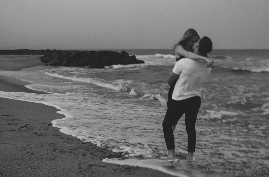 couple on the beach standing at the edge of the water while the man holds his partner in his arms while kissing